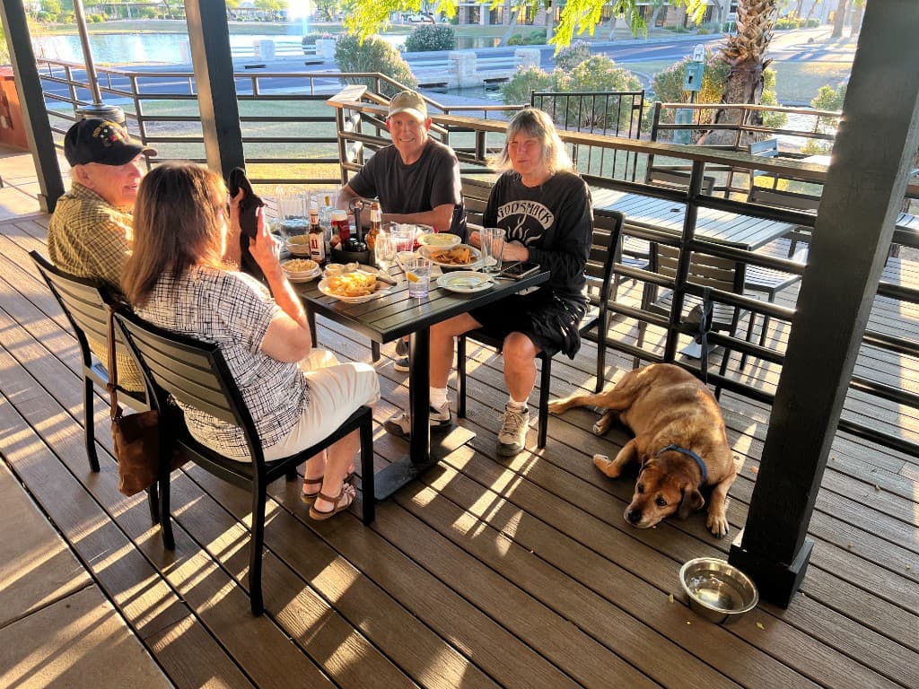 Guests dining on The Grove patio with a dog nearby and a lake view beyond